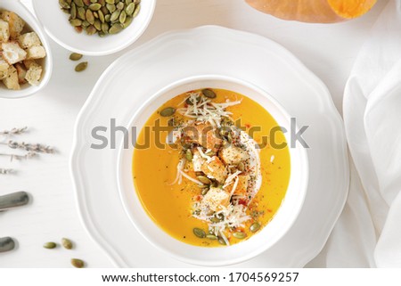 Pumpkin soup, a piece of pumpkin, seeds and crackers lie on a white table. Nearby is a white towel and serving utensils.
Close up.