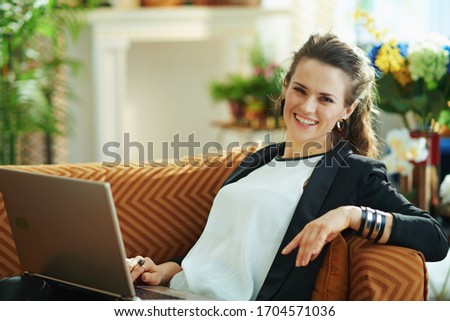 smiling modern woman in white blouse and black jacket in the modern living room in sunny day editing on a laptop while sitting on divan.