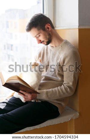 A young man sits on a windowsill and reads a book. Next to the man sits a white cat with red spots. A man is wearing a beige sweater.