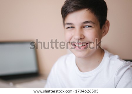 Happy teenage boy sits by the table and study at home. Boy study online during quarantine. Background of laptop and books