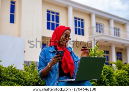 young black beautiful lady sitting out and working on her laptop while making online payment with her card on her phone