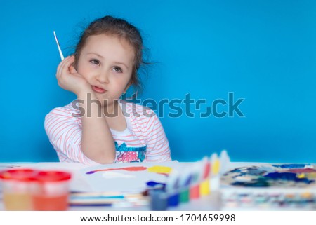 A cute child with a brush in his hands looks up thoughtfully. The girl draws with colored paints on a white sheet, the foreground is blurred. Copy space. Mock up