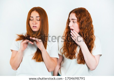 two redheaded young women standing on isolated white backgroung one is checking her split ends while another looks upset, style and beauty concept