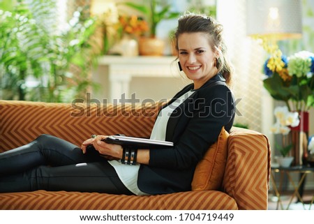 smiling modern housewife in white blouse and black jacket with closed laptop sitting on couch at modern home in sunny day.