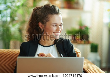 happy stylish female in white blouse and black jacket in the modern living room in sunny day using laptop while sitting on sofa.