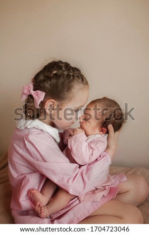 A portrait of two sisters in pink dresses with white collars hugging. The elder sister holds a newborn baby girl gently, trying to kiss her. Loving family lifestyle picture.