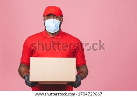 Delivery african american man employee in red cap blank t-shirt uniform face mask gloves hold empty cardboard box isolated on pink background. Service quarantine pandemic coronavirus virus 2019-ncov.  Royalty-Free Stock Photo #1704729667