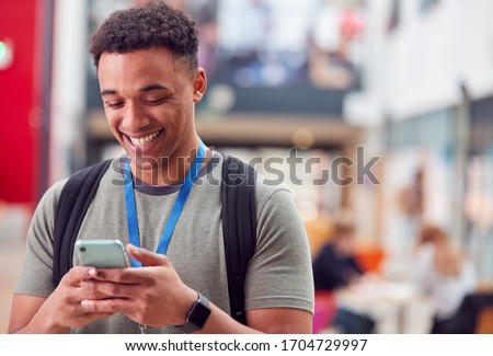 Smiling Male College Student Checking Mobile Phone In Busy Communal Campus Building Royalty-Free Stock Photo #1704729997
