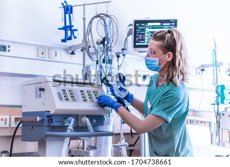 COVID19/2019-ncov concept: nurse, wearing a surgical mask, checks the settings of a mechanical ventilation machine, which is seen in the foreground. therapy used for lung breathing, in intensive care Royalty-Free Stock Photo #1704738661
