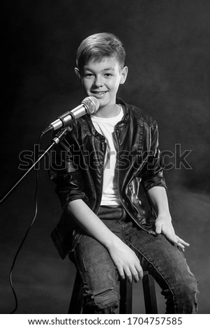 Monochrome portrait of caucasian teenager in white t-shirt, blue jeans and leather jacket with microphone singing on dark background. Hobby, popularity and glory concept