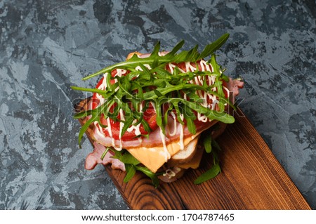 Very tasty big sandwich with ham or balyk, arugula, cheese, tomatoes and mayonnaise on a cutting board in rustic style on a gray background view from the top, copy space