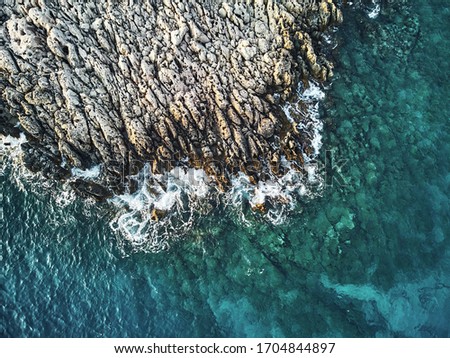 top drone view of a stony coast clashing into the blue ocean sea. Structure and Pattern in artistic way of the sharp rocks in the water. Aerial top shot on warm summer waves.