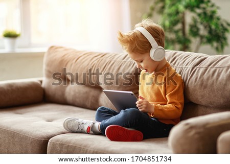 Full body delighted little boy in headphones smiling and watching interesting cartoon on tablet while sitting crossed legged on sofa at home
