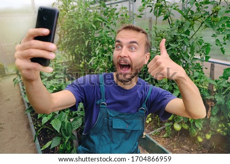 Funny millennial farmer making selfie on smartphone in his greenhouse, showing like sign, because harvest is good. Organic farming, local bio farm, sustainable food concept.