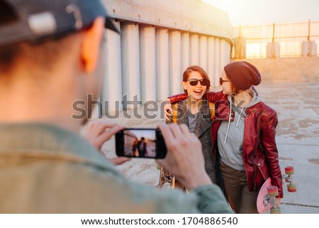 Man taking pictures of two young happy smiling women with short hair in sunglasses with skateboards. Sunset in a city. Gray floor and building.