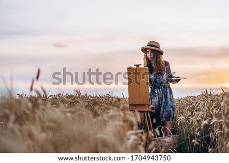 Portrait of smiling female artist with curly hair in hat. Girl draws a picture of a landscape in a wheat field. Copy space.