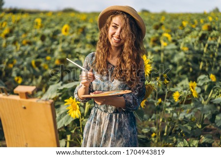 A young woman with curly hair and wearing a hat is painting in nature. A woman stands in a sunflower field on a beautiful day.
