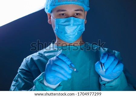 close-up portrait of a caucasian doctor surgeon, in a sterile suit, mask, holding a scalpel and needle holder with a needle, guiding them to the operating wound, getting ready to operate.