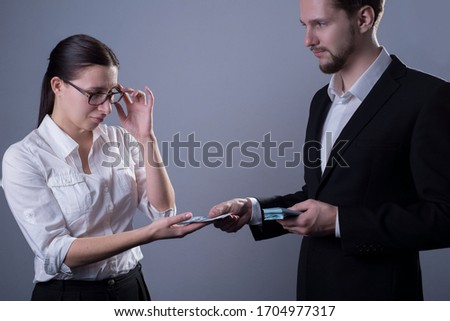 Portrait of two young businessmen. A business woman takes off her glasses, looks sadly at the small salary that her boss puts into her hand. Studio photo on a gray background.