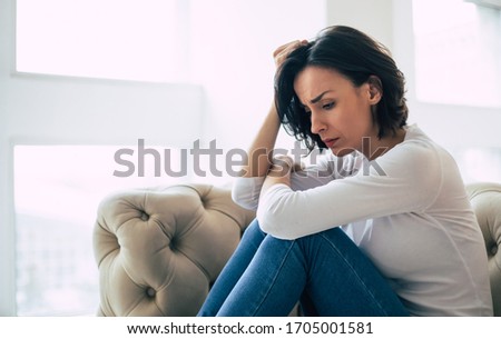 Empty and numb. Close-up photo of a depressive girl who is hugging her knees and touching her head while sitting near the window.