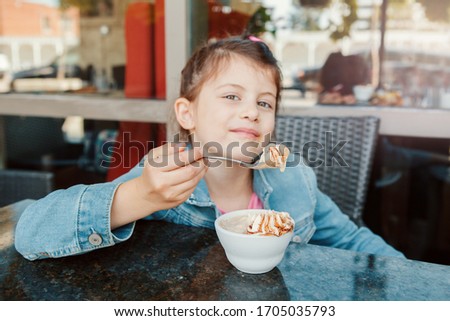 Funny Caucasian preschool girl eating sweet dessert with spoon in cafe. Child kid having fun in restaurant patio enjoying food drink. Happy authentic childhood lifestyle. 