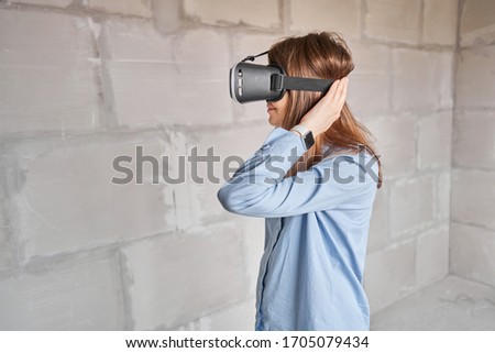 Woman architect or construction engineer wear virtual reality glasses inside a building site. Concrete walls in new apartment, concept of repair.