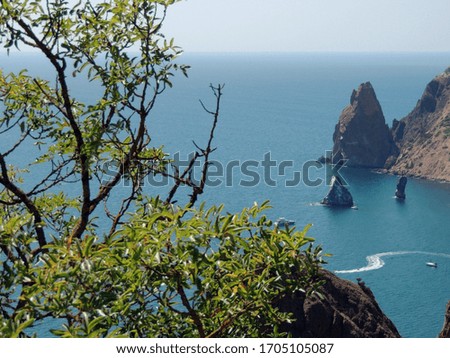 Photo of rocks and cape Fiolent near Sevastopol in the republic of Crimea. Sea, boat and rocks photographed through trees with green leaves.
