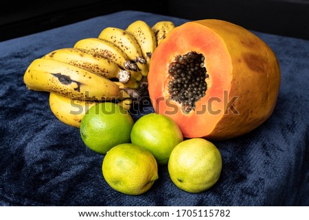 Close up to a fresh sliced orange papaya with seeds inside, a freckled bananas cluster and three green lemons over a blue navy and black background. Fruit and eating concept photography