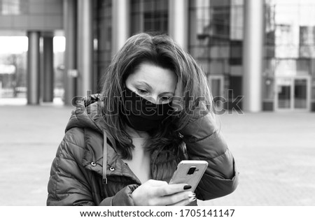 black and white photo of a woman with a black mask on her face against background of business buildings, holding her head and looking at the screen of a smartphone, bad news about the epidemic.