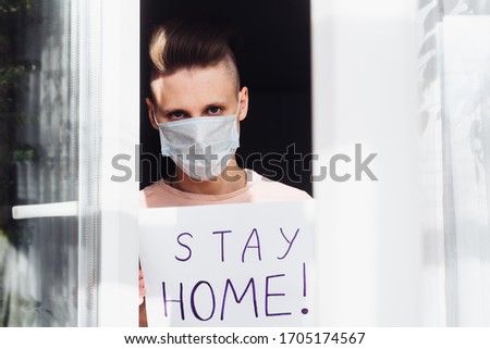 A young man in a protective mask stands at home outside the window and shows a table with the words Stay at home. A call for self-isolation due to a pandemic and the risk of coronavirus infection