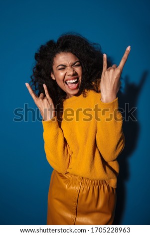 Photo of happy young african woman posing isolated over blue wall background showing rock gesture.