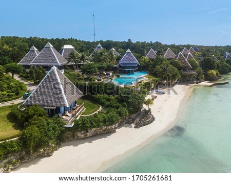 Aerial of resort bungalows and a pool, atop a short cliff and beside a white sand beach and tropical waters during a clear day.