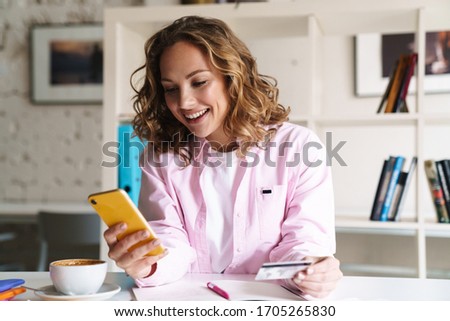 Photo of smiling nice woman using cellphone and holding credit card while drinking coffee in cafe