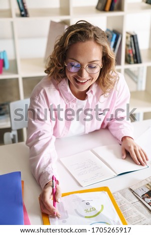 Photo of joyful blonde woman wearing eyeglasses smiling and doing homework while sitting in room