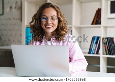 Photo of joyful blonde woman wearing eyeglasses smiling and using laptop while sitting in cafe