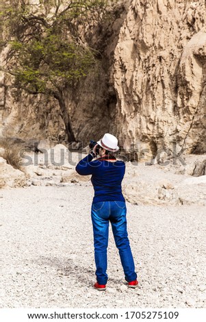 Slender elderly woman in jeans and a hat photographs the canyon. Stone canyon in the mountains of the Judean desert. The coast of the famous Dead Sea. Ecological, active and photo tourism concept