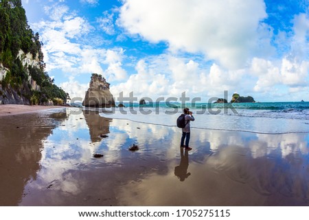 Travel to New Zealand. Ocean tide in the Cathedral Cove. Woman photographs the ocean. Mirror reflections of clouds in wet sand. The concept of exotic, ecological and photo tourism