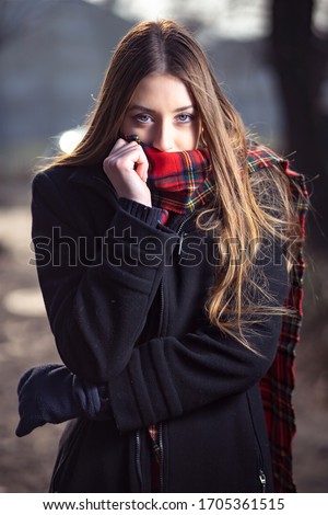 Outdoor portrait photo of cute blond girl in black jacket and red scarf looking like she is cold