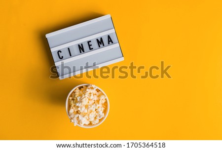 Popcorn in a vase with text CINEMA on white lightbox isolated on yellow background. Flat lay banner, top view. To go to the cinema.