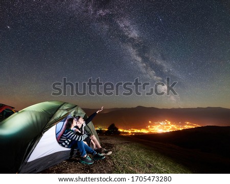Romantic couple travellers having a rest in glowing tourist tent. Man pointing at amazing night sky full of stars and Milky way. On the background beautiful starry sky, mountains and luminous city