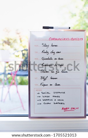 weekly planning sign with stay at home in different languages for each day, indoors on window with small pink table and 2 chairs on terrace on background. Sun is shining