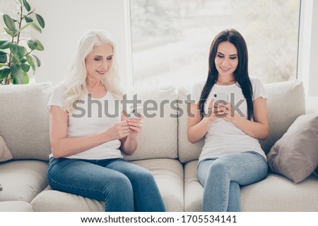 Portrait of two nice attractive lovely pretty cheerful focused women granddaughter grandmother using digital device sitting on divan in light white interior room house flat apartment