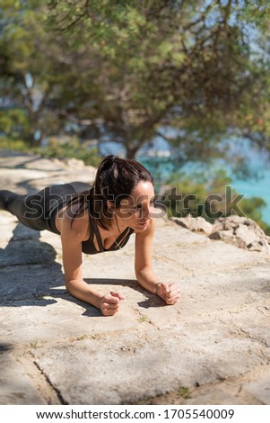 Attractive young brunette woman with tan skin doing a plank in front of the beach on a summer day. Lifestyle workout, exercising hard. Being active. Sport outside, CrossFit