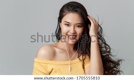 Close up young attractive Asian woman wearing yellow t shirt, earphones listening music with smile face, studio head shot isolated on grey background.