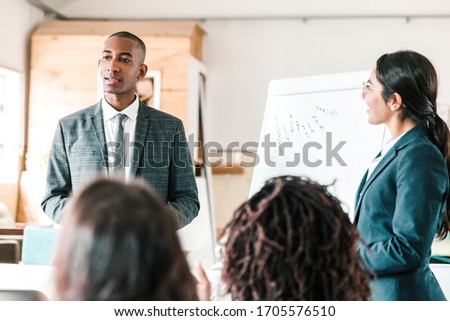 Cheerful young speakers standing near whiteboard. Back view of employees sitting and listening colleagues. Business meeting concept