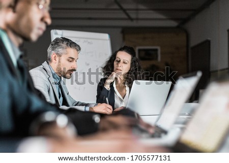 Confident people working over new project in dark office. Thoughtful business team sitting at long table with laptops. Teamwork, working late concept