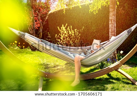 Young relaxed girl reading book in hammock in garden at home at bright sunset. Slow living, gadget detox and weekend leisure activity. Quarantine and self isolation period Royalty-Free Stock Photo #1705606171