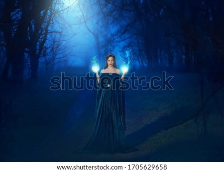 Mystic beauty woman evil witch in dark night fog forest blue moon light. Hands burning magic spell fire. Vampire queen demon power face. Vintage medieval dress cape. Scary Lady Halloween style clothes