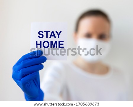 Woman with wearing medical face mask holding white sticker with words STAY HOME on white background