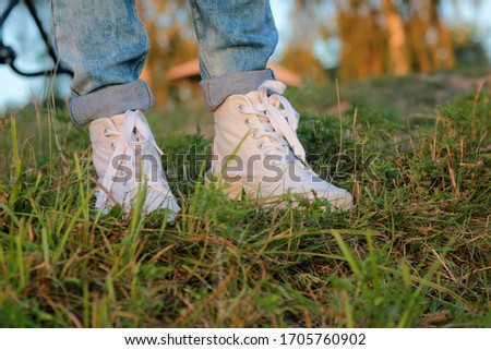 Close up of teenager's feet in modern and trendy white sneakers and rolled up jeans. Sunny summer day, green grass background. 